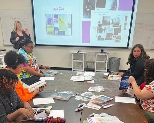  Students sit at table covered in art supplies in front of presenter.